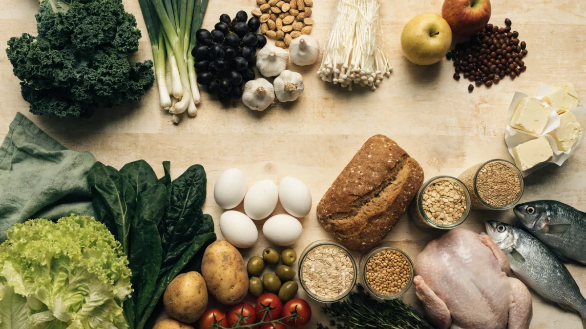Bogaty wybór świeżych produktów spożywczych — owoce, jagody, warzywa, jajka, pieczywo, ryby i kasze — ułożony na drewnianym stole, widok z góry.